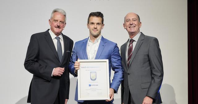 Drei Männer in Anzügen vor einem weißen Hintergrund, der mittlere Mann hält eine Urkunde in der Hand.