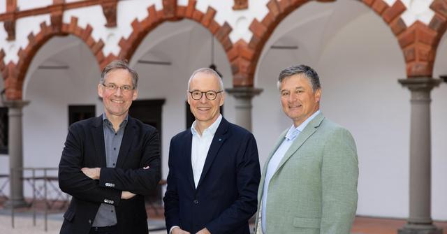 Drei Männer mittleren Alters stehen in einem Innenhof.