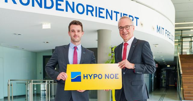 Zwei Männer in Anzug und Krawatte halten ein Schild mit gelben Hintergrund und blauer Aufschrift "HYPO NOE".