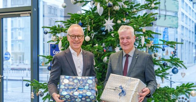 Zwei Männer in Anzug stehen vor einem geschmückten Christbaum und halten je ein Geschenkspackerl in der Hand.