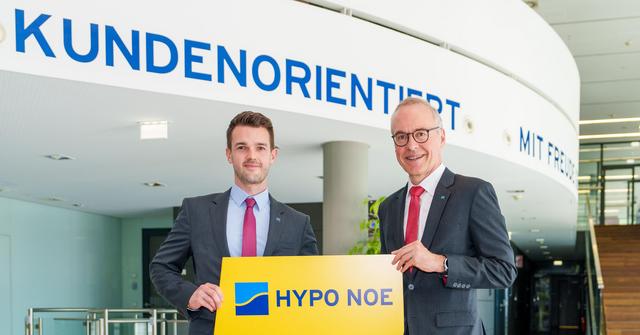 Zwei Männer mittleren Alters im Anzug mit Krawatte stehen im Eingangsbereich eines Bürogebäudes und halten ein Schild.