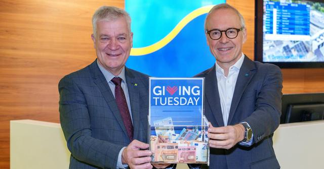 Zwei Männer im Anzug halten gemeinsam vor sich eine Spendenbox, die mit Geld gefüllt ist.