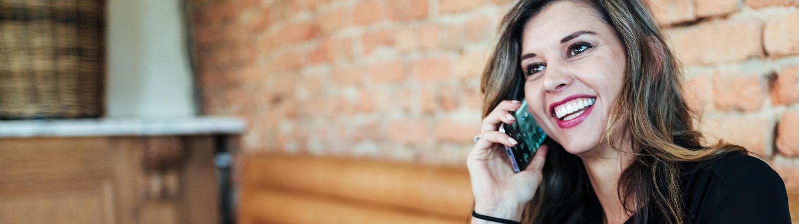 Junge Frau sitzt lächelnd vor ihrem Laptop und drückt das Handy ans Ohr. Sie nutzt die Kontaktmöglichkeiten zur HYPO NOE