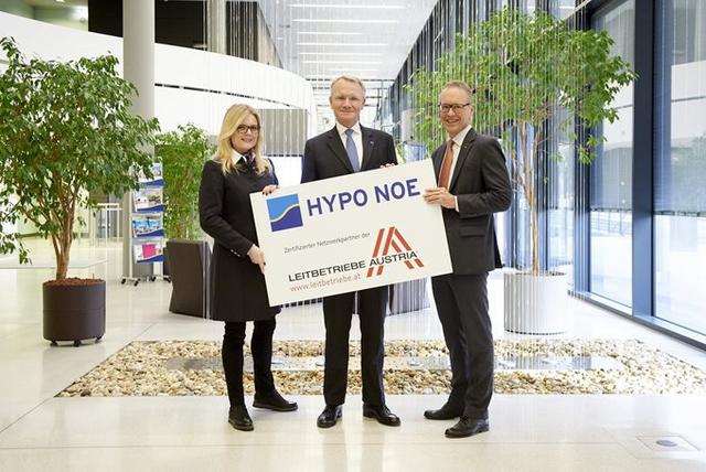Eine Frau und zwei Männer in Businesskleidung halten ein Schild mit dem HYPO NOE Logo und dem Leitbetriebe Logo