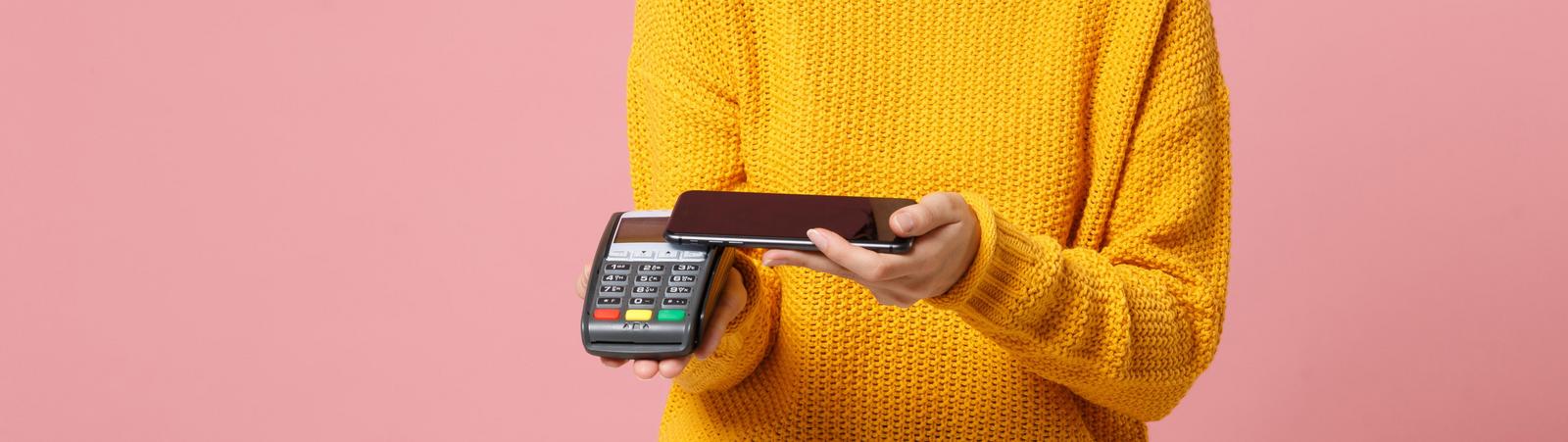 Smiling young woman in yellow sweater posing isolated on pink background. People lifestyle concept. Mock up copy space. Hold payment terminal to process and acquire credit card payments, mobile phone.