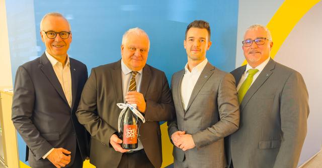 Vier Männer in Anzug stehen nebeneinander, einer hält eine Weinflasche als Abschiedsgeschenk in der Hand.