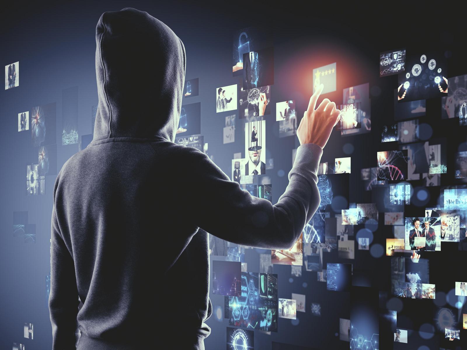 Eine Person mit Kapuzenhoodie steht in Dunkelheit vor einer digitalen Wandtafel mit Internetsymbolen